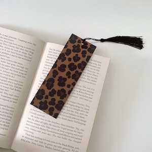 Puede incluir: Un marcapáginas de color marrón y negro con estampado de leopardo y un borla negra. El marcapáginas está hecho de un material suave y texturizado.