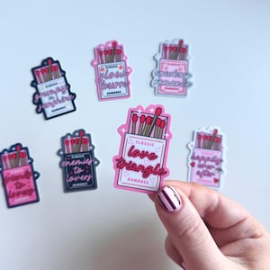 May include: A set of ten pink and white matchbox stickers with different romantic phrases. The stickers feature a box of matches with red matchsticks and the text "Classic Romance" on the box. The phrases on the stickers include "Grumpy Sunshine", "Glow Burn", "Cowboy Romance", "Enemies to Lovers", "Happily Ever After", and "Classic Love Triangle".
