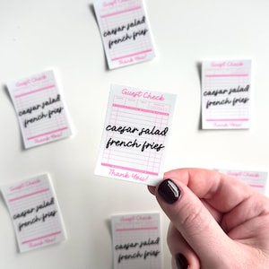 May include: A set of six pink and white guest checks with the text "caesar salad french fries" written in black cursive. The checks are on a white background.
