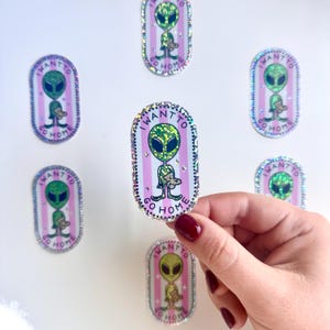 May include: Six oval stickers with a pink and white striped background. Each sticker features a green alien holding a pink teddy bear. The text on the stickers reads "I WANT TO GO HOME".