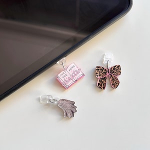 May include: Three decorative phone charms. One is a pink book with the text "Just One More Chapter". Another is a bow with a leopard print pattern and pink accents. The third is a silver wing.