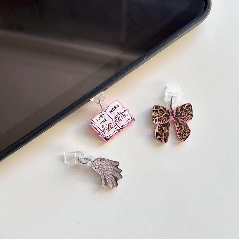 May include: Three decorative phone charms. One is a pink book with the text "Just One More Chapter". Another is a bow with a leopard print pattern and pink accents. The third is a silver wing.