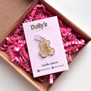 May include: A clear Kindle charm featuring a brown teddy bear design, presented on a pink card with the text "Dolly's doodles" and "kindle charm", nestled in a brown box with pink paper.