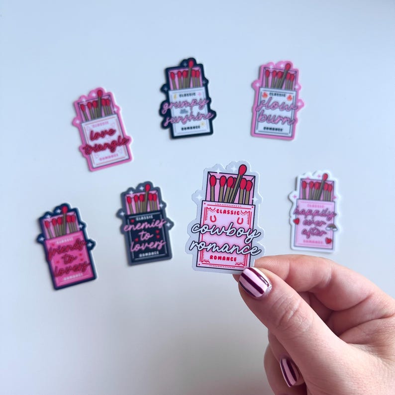 May include: A set of nine matchbox-style stickers with various romantic themes. Each sticker features a pink and white matchbox with red matchsticks and a pink and white text label. The labels include "love triangle", "grumpy sunshine", "classic u cowboy romance", "enemies to lovers", "glow burn", "happily ever after", "friends to lovers".