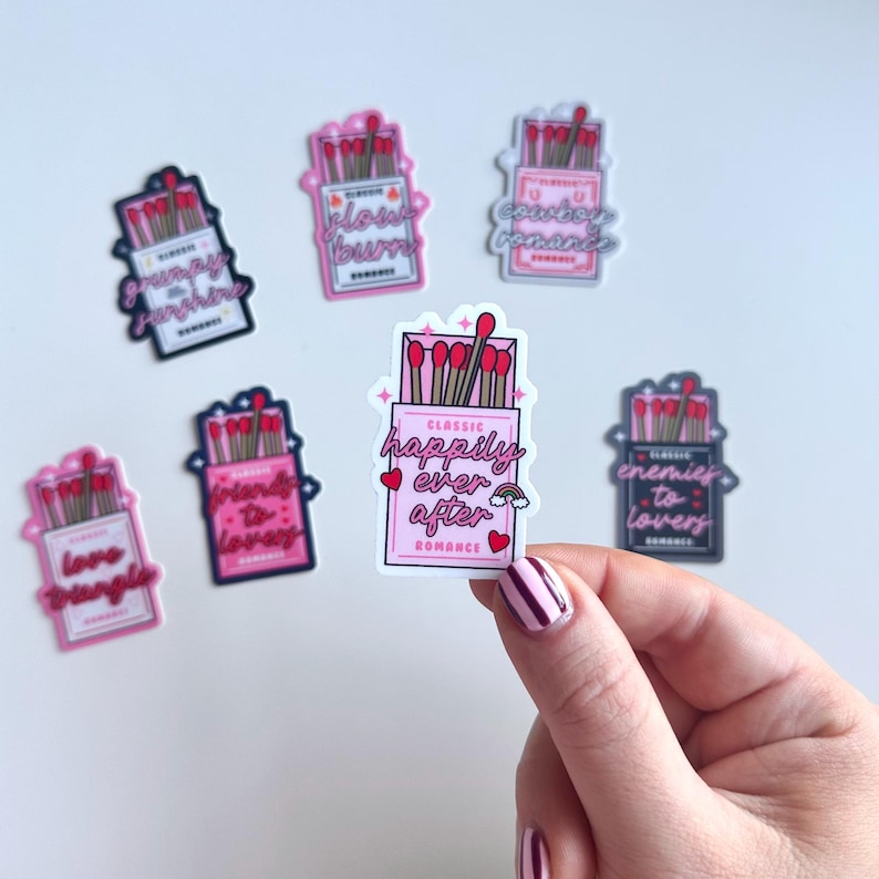 May include: A set of ten pink and black matchbox stickers with different romantic phrases. The stickers feature a matchbox with a red and white matchstick inside. The phrases include "Grumpy Sunshine", "Slow Burn", "Cowboy Romance", "Friends to Lovers", "Love Triangle", "Happily Ever After", "Enemies to Lovers", and "Classic Romance".