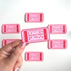 May include: Pink and red ticket with the text "LIFETIME MEMBERSHIP TO THE Romance Book Club SPARKS GUARANTEED" and "ADMITS ONE 0003". The ticket is being held by a hand with pink nail polish.