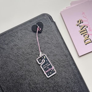 Puede incluir: Un dije negro en forma de coraz&oacute;n con una cuerda rosa adjunta a una etiqueta rectangular que dice "DO NOT DISTURB I'M reading" con un dise&ntilde;o de luna creciente y estrellas. La etiqueta cuelga de una superficie negra texturizada.