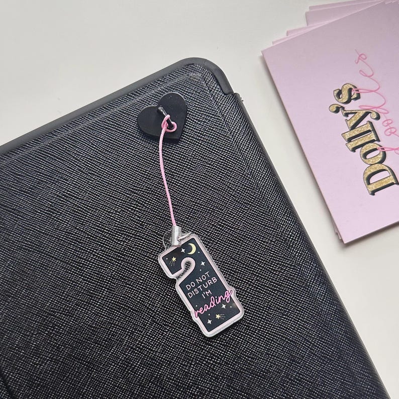 Puede incluir: Un dije negro en forma de coraz&oacute;n con una cuerda rosa adjunta a una etiqueta rectangular que dice "DO NOT DISTURB I'M reading" con un dise&ntilde;o de luna creciente y estrellas. La etiqueta cuelga de una superficie negra texturizada.