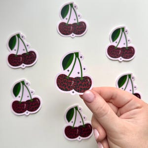 May include: Cherry stickers with a leopard print design. Each sticker features two cherries in a deep red hue with a black leopard pattern, green leaves, and a light pink border. The stickers are scattered on a white surface.