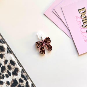 May include: A bow-shaped earring with a pink center and a leopard print design. The earring is attached to a clear earring post. Pink cards with gold text are in the background. A leopard print pattern is visible on the left.