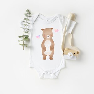 May include: White baby onesie with a brown bear illustration and pink hearts. The onesie is on a white background.
