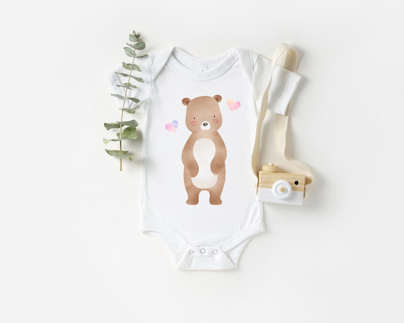 May include: White baby onesie with a brown bear illustration and pink hearts. The onesie is on a white background.