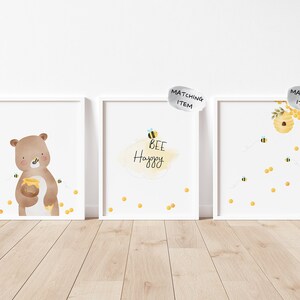 May include: Three white framed prints with a bee theme. The first print features a brown bear holding a jar of honey with bees flying around it. The second print has the text "Bee Happy" with a bee and honeycomb design. The third print has a honeycomb with bees flying around it.