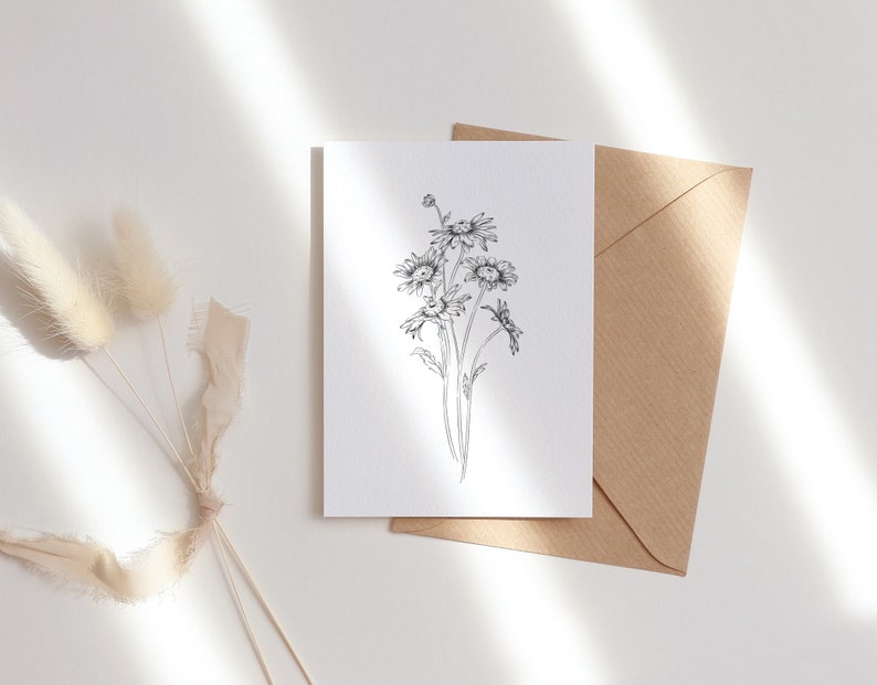 May include: A white card with a black and white line drawing of a bouquet of daisies. The card is sitting on a brown envelope.