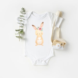 May include: White baby onesie with a brown bunny wearing a pink flower crown. The bunny is surrounded by green leaves.