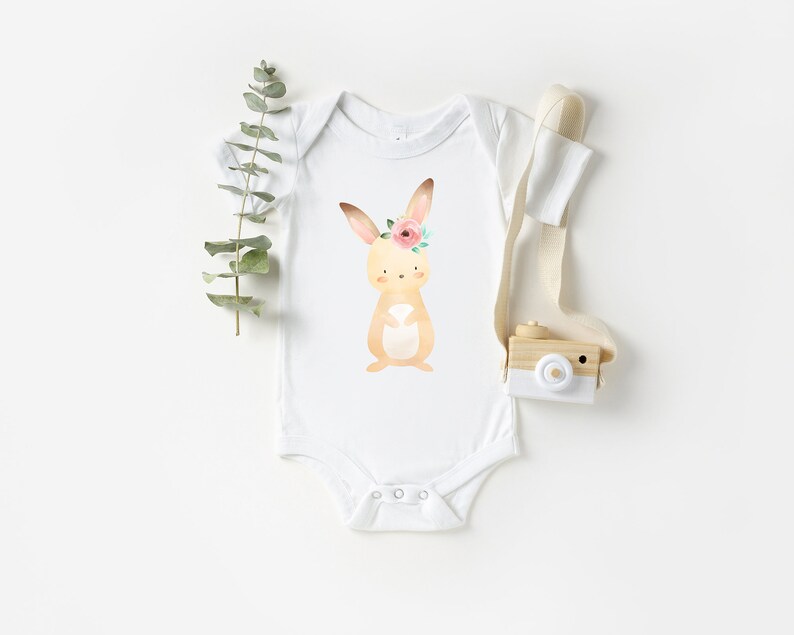 May include: White baby onesie with a brown bunny wearing a pink flower crown. The bunny is surrounded by green leaves.