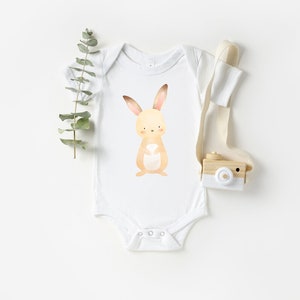 May include: A white baby onesie with a brown bunny rabbit illustration on the front. The bunny has a white belly and pink ears.
