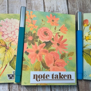 May include: Three floral patterned notebooks with silver metal pens. The notebooks have a green background with pink, yellow, and white flowers. The pens are teal, pink, and blue. The middle notebook has the text "note taken" on the front.