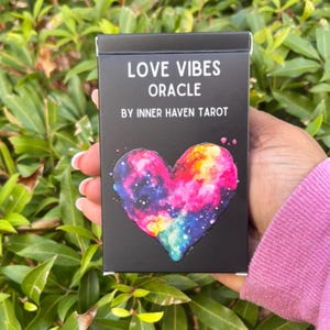 May include: A black oracle card box with the text "LOVE VIBES ORACLE BY INNER HAVEN TAROT." The box features a colorful heart design with a galaxy pattern in pink, blue, yellow, and purple. The box is held against a background of green foliage.