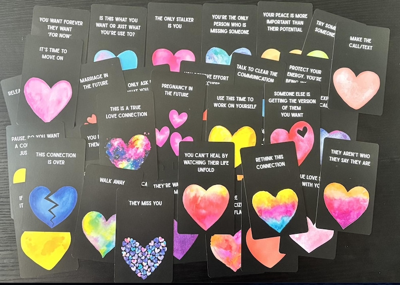 May include: A selection of black tarot cards, each with a colourful heart design and a different phrase. The cards display pink, blue, yellow, and purple hearts, with text such as "Marriage in the Future" and "This is a True Love Connection."