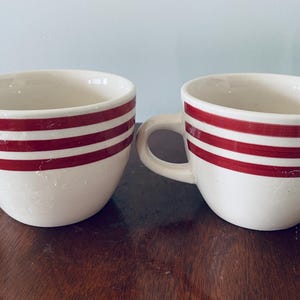 May include: Two white ceramic mugs with red horizontal stripes. Each mug has a rounded shape, a handle, and a glossy finish. The mugs are placed on a dark wooden surface, with a light blue background.
