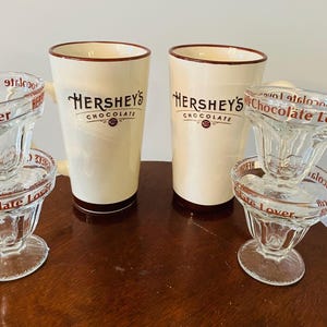 May include: Two Hershey's chocolate mugs and two glass sundae dishes. The mugs are cream-colored with the Hershey's logo in black and brown. The sundae dishes are clear glass with "Chocolate Lover" printed in red.