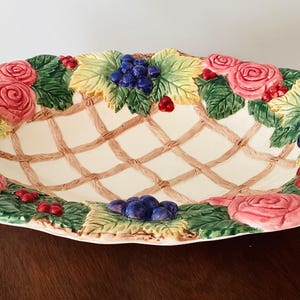 May include: A white ceramic serving bowl with a woven lattice design. The bowl is decorated with sculpted roses, grapes, and leaves in shades of pink, green, yellow, and purple.