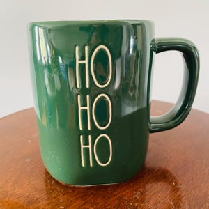 Puede incluir: Una taza de cerámica verde oscuro con un asa curvada. La taza presenta la palabra "HO" repetida tres veces en una fuente blanca en relieve. La taza está sobre una superficie de madera.