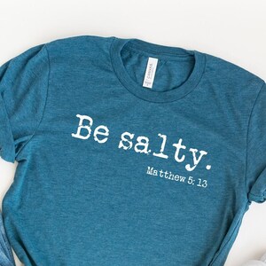 May include: A teal blue t-shirt with the text "Be salty. Matthew 5:13" in white. The shirt is laid out on a white surface with a pair of blue jeans and white sneakers.