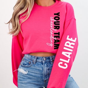 May include: A pink cropped sweatshirt with long sleeves. The left sleeve has the text "CLAIRE YOUR TEAM cheerleading" in white and black lettering.