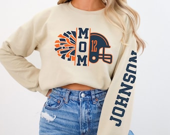May include: A beige sweatshirt with a graphic of a football helmet and a cheerleading pom-pom. The text "MOM" is printed on the graphic. The text "JOHNSON" is printed on the sleeve.