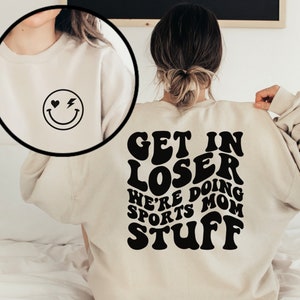 K&ouml;nnte beinhalten: Ein beiger Pullover mit einem schwarzen Aufdruck auf der R&uuml;ckseite, der in einer Retro-Schrift "Get in Loser We're Doing Sports Mom Stuff" lautet. Die Vorderseite des Pullovers hat ein kleines schwarzes Smiley mit einem Herz und einem Blitz.