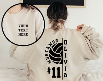 Może przedstawiać: Beżowa bluza z czarnym nadrukiem piłki siatkowej, tekstem "MUSTANGS #11 OLIVIA" i czarną piłką siatkową.