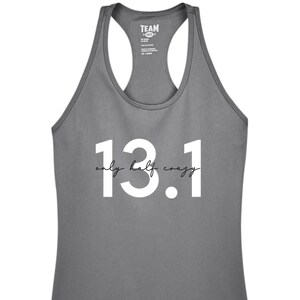 May include: Gray racerback tank top with the text "13.1 only half crazy" in white. The tank top has a scoop neck and is made of a lightweight material. The text is in a bold, sans-serif font with the words "only half crazy" in a cursive font.
