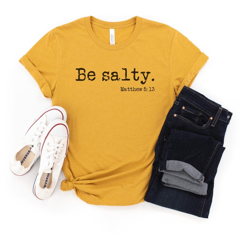 May include: A mustard yellow t-shirt with the text "Be salty. Matthew 5:13" printed on the front. The shirt is folded and laid flat on a white background.