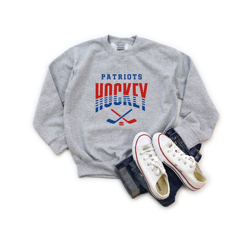 Puede incluir: Sudadera gris con el texto "Patriots Hockey" en letras rojas y azules. El texto est&aacute; rodeado por un dise&ntilde;o a rayas rojas y azules. Dos palos de hockey cruzados y un disco se encuentran debajo del texto.