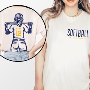 Puede incluir: Camiseta color crema con la frase "SOFTBALL aunt" en azul marino y amarillo. La parte trasera de la camiseta presenta un gr&aacute;fico de jugadora de softbol con un bate, el n&uacute;mero 12 y espacio para un nombre. El dise&ntilde;o es en azul marino y amarillo.