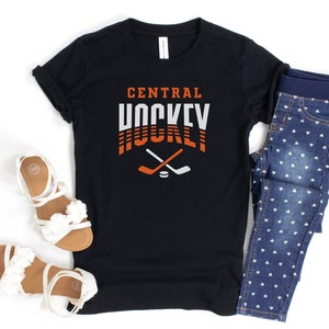 Puede incluir: Camiseta negra con el texto "CENTRAL HOCKEY" en naranja y blanco con un gr&aacute;fico de palo de hockey. La camiseta se combina con jeans azules con patrones de corazones blancos.
