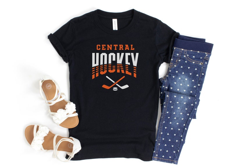 Puede incluir: Camiseta negra con el texto "CENTRAL HOCKEY" en naranja y blanco con un gr&aacute;fico de palo de hockey. La camiseta se combina con jeans azules con patrones de corazones blancos.