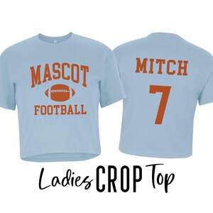 Pu&ograve; includere: Top corto azzurro con le parole "MASCOT FOOTBALL" e una grafica di football americano arancione. La parte posteriore della maglietta ha il nome "MITCH" e il numero "7" in arancione. Le parole "Ladies CROP Top" sono scritte in nero.