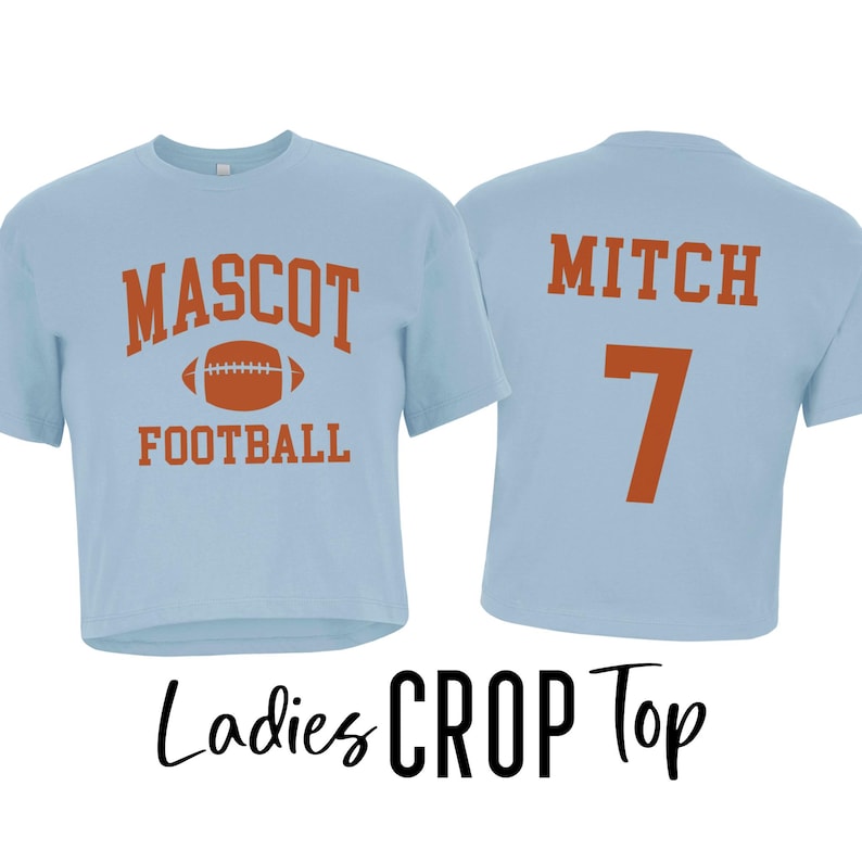 Pu&ograve; includere: Top corto azzurro con le parole "MASCOT FOOTBALL" e una grafica di football americano arancione. La parte posteriore della maglietta ha il nome "MITCH" e il numero "7" in arancione. Le parole "Ladies CROP Top" sono scritte in nero.