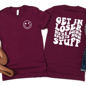 K&ouml;nnte beinhalten: Burgunderfarbenes T-Shirt mit einem wei&szlig;en Smiley auf der Vorderseite und dem Text "Get in loser we're doing sports mom stuff" in einer Retro-Schriftart auf der R&uuml;ckseite.