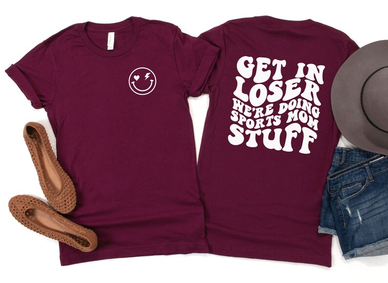 K&ouml;nnte beinhalten: Burgunderfarbenes T-Shirt mit einem wei&szlig;en Smiley auf der Vorderseite und dem Text "Get in loser we're doing sports mom stuff" in einer Retro-Schriftart auf der R&uuml;ckseite.