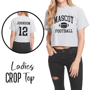 Pu&ograve; includere: Un crop top grigio chiaro con la scritta "MASCOT" sopra una grafica di football americano e la parola "FOOTBALL" sotto. La parte posteriore della maglietta ha il nome "JOHNSON" e il numero "12" in nero. Sono visibili anche le parole "Ladies CROP Top".
