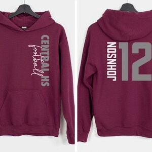 May include: Burgundy hoodie with a front pocket and hood. The front features the text "CENTRAL HS football" in white and gray. The back displays "JOHNSON" and the number "12" in gray.