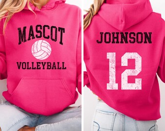 Puede incluir: Una sudadera con capucha rosa brillante con la palabra "MASCOT" encima de un gráfico de voleibol y la palabra "VOLLEYBALL" debajo. La parte trasera muestra el nombre "JOHNSON" y el número "12" en blanco.