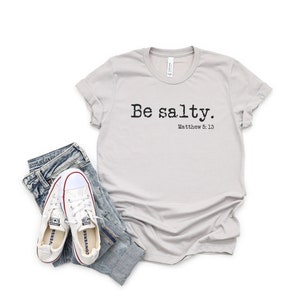 May include: A light gray t-shirt with the text "Be salty. Matthew 5:13" printed in black. The shirt is laying flat on a white background with a pair of white sneakers and blue jeans.