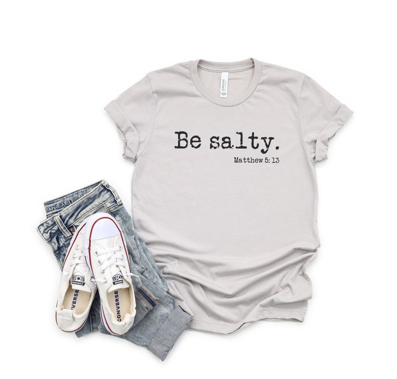 May include: A light gray t-shirt with the text "Be salty. Matthew 5:13" printed in black. The shirt is laying flat on a white background with a pair of white sneakers and blue jeans.