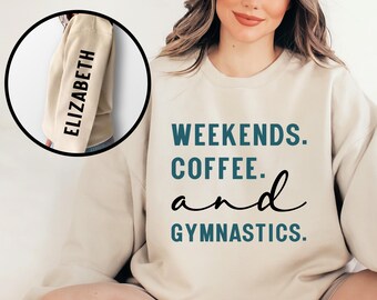 May include: A beige sweatshirt with the words "WEEKENDS. COFFEE. and GYMNASTICS." in teal and black script. The left sleeve in a circular inset shows the name "ELIZABETH" in black vertical text. A casual, comfortable top.