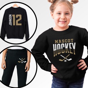 K&ouml;nnte beinhalten: Schwarzer Pullover mit dem Text "MASCOT HOCKEY" in goldenen und wei&szlig;en Buchstaben. Der Pullover hat ein Design mit gekreuzten Hockeyschl&auml;gern und einem Puck. Ein schwarzer Pullover mit der Nummer "12" und dem Namen "JOHNSON" in goldenen Buchstaben. Schwarze Jogginghose mit einem Hockeyschl&auml;ger- und Puck-Design in Gold.